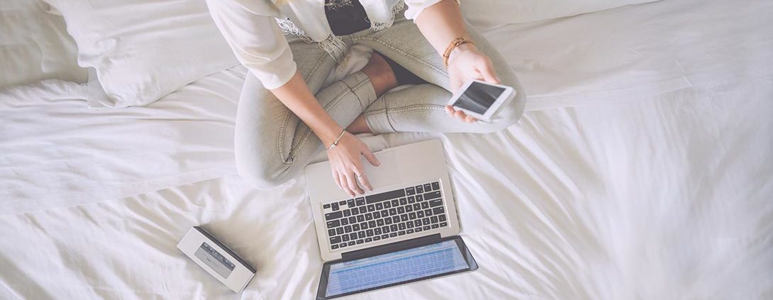 Frau mit Smartphone und Laptop sitzt auf Bett